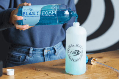 Bottle of pH Neutral Snow Foaming Soap being poured into a Snow Foaming Applicator Gun in front of the Wat-er Blast Logo